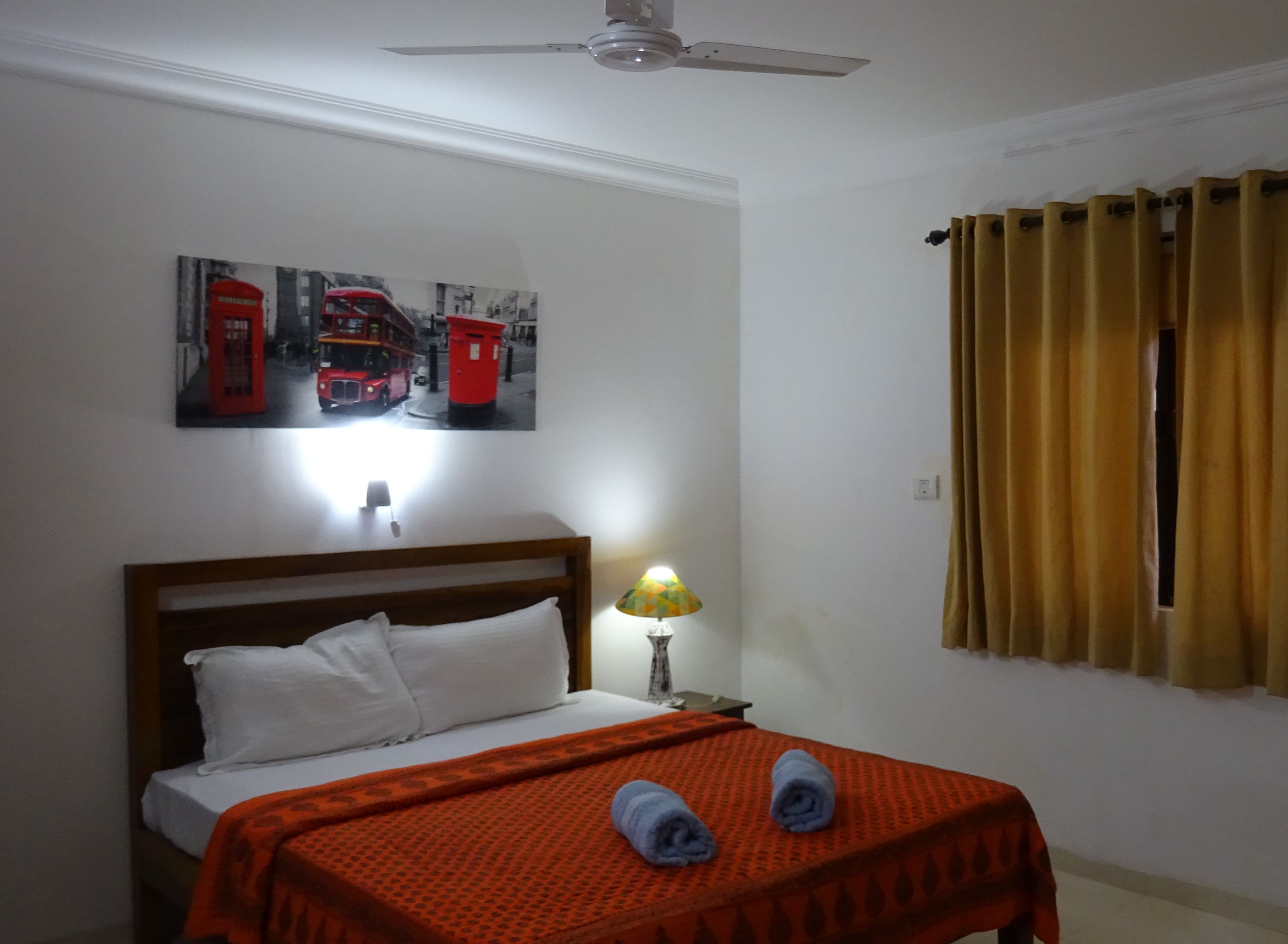 A neatly made bed with a red blanket, a lamp on a bedside table, and a wall with a red telephone booth picture.