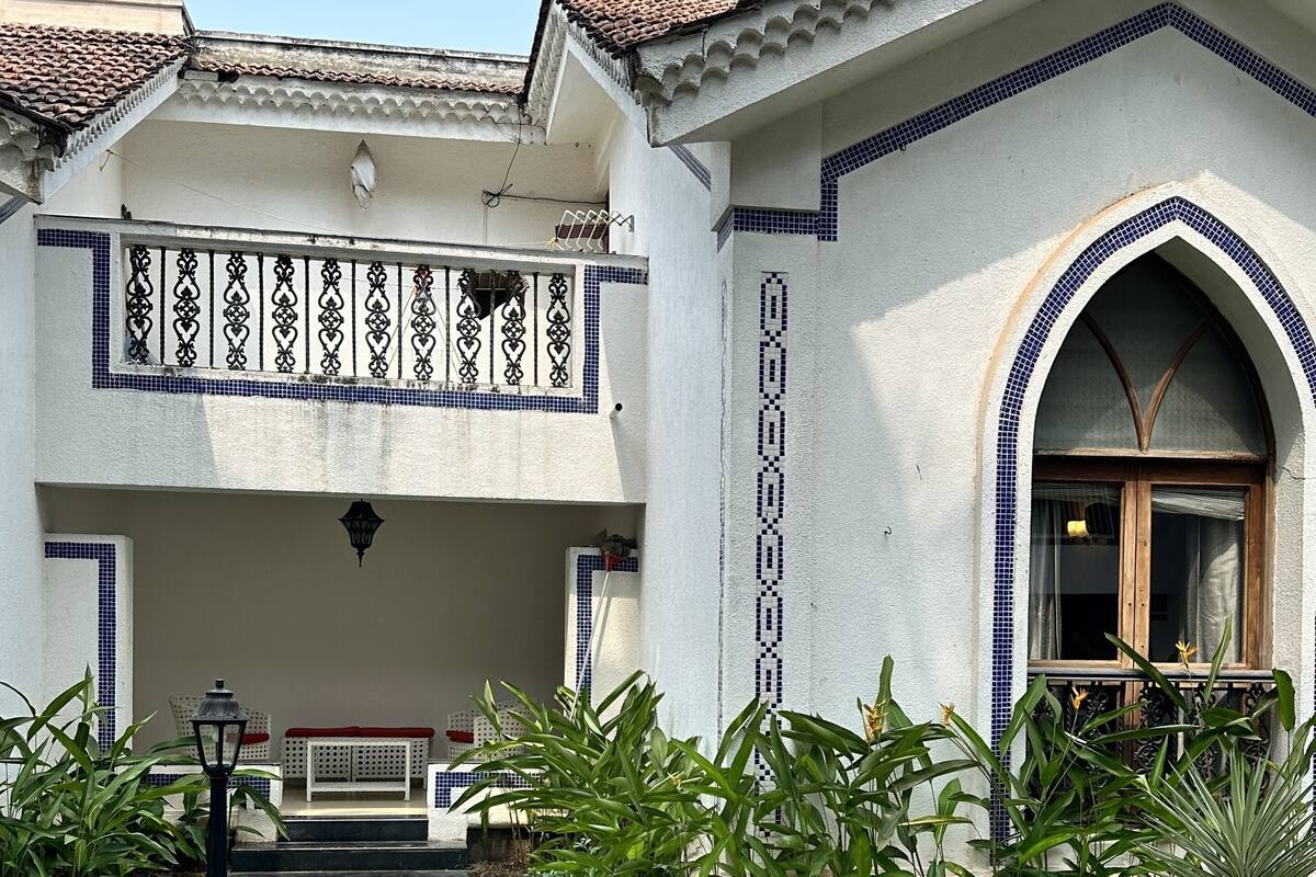 Un edificio de dos pisos con techo de tejas, un balcón con barandillas decorativas y un jardín con diversas plantas.