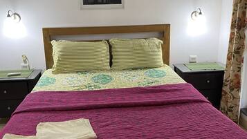 A bedroom with a bed, two bedside tables, an air conditioner, a wall-mounted lamp, and a window with a view of trees.