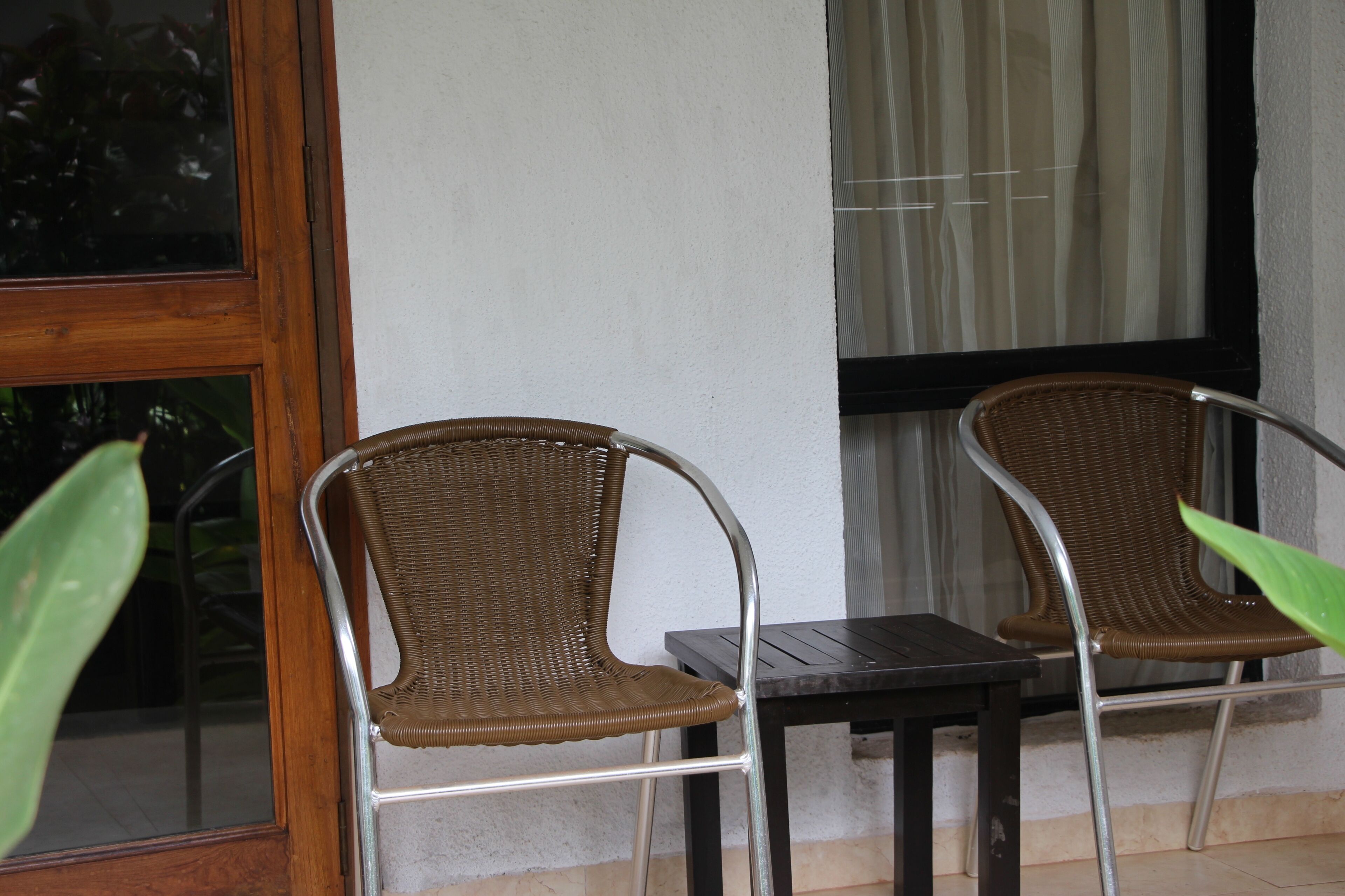 Two wicker chairs and a small table outside a building with a window and a curtain.