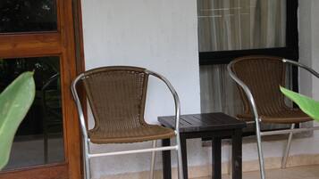 Two wicker chairs and a small table outside a building with a window and a curtain.