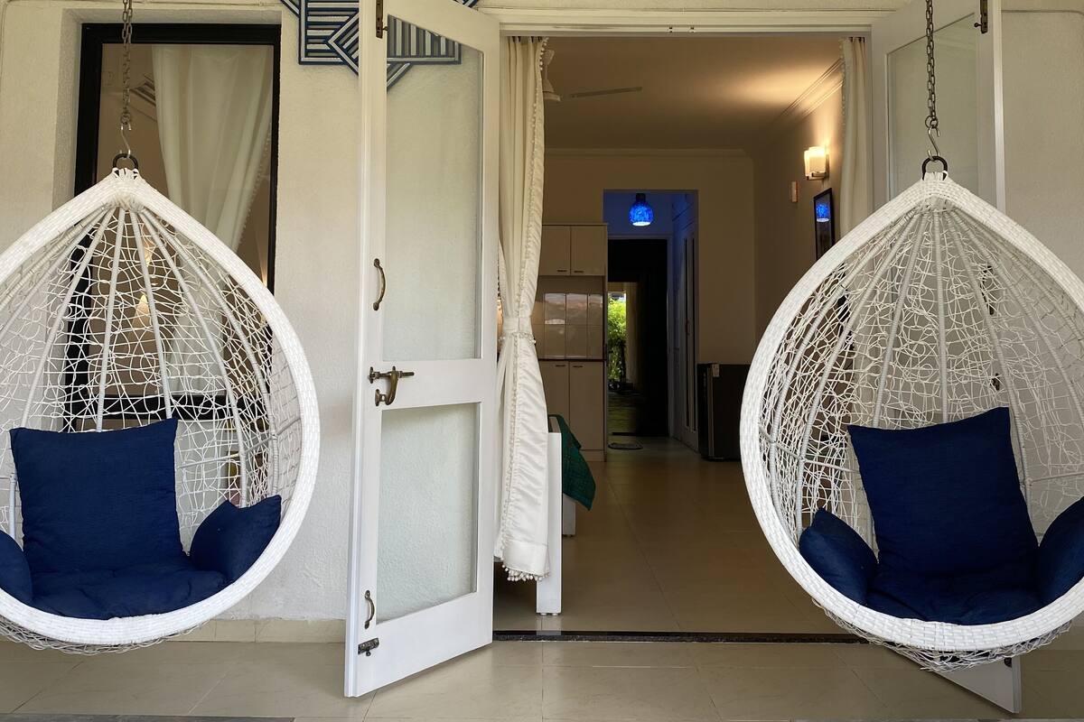Una habitación con dos sillas colgantes blancas con cojines azules, una puerta blanca y un ventilador de techo.