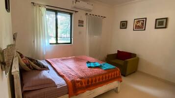 Un dormitorio con cama, una mesita, una silla, un ventilador de techo y una ventana con cortinas.