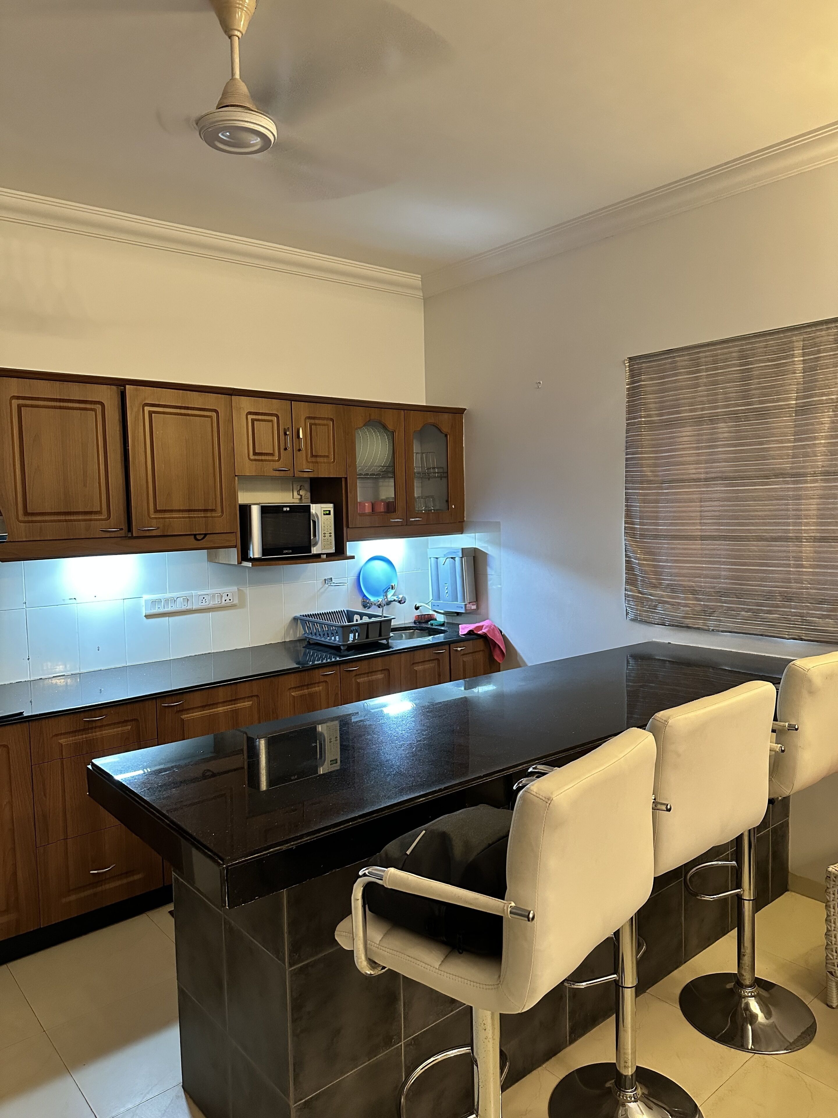 A kitchen with wooden cabinets, a black countertop, a microwave, and a sink. There are two white bar stools.