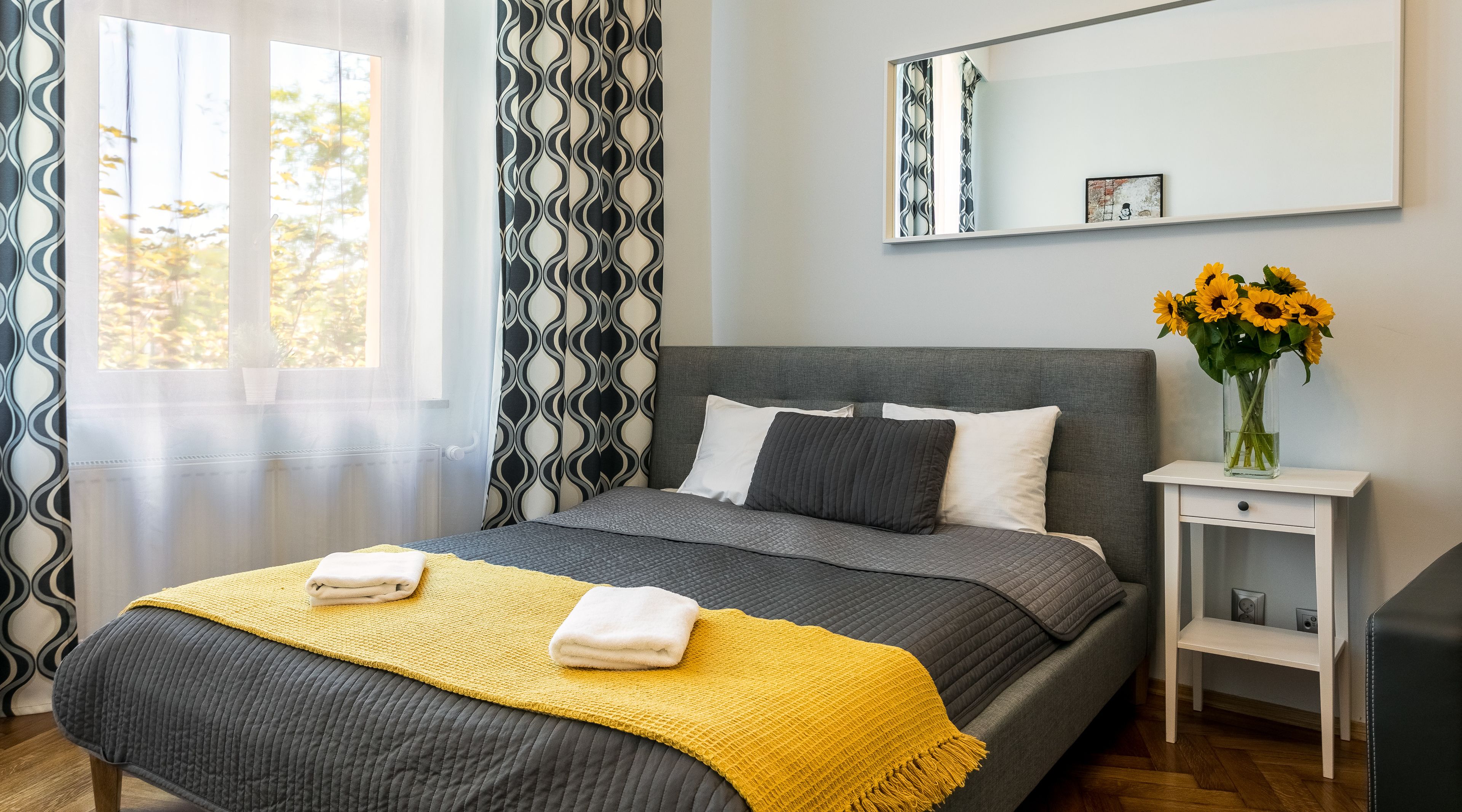 Ein modernes Schlafzimmer mit einem grauen Bett, weißen Kissen, einer gelben Tagesdecke und einem Nachtisch mit einer Vase voller Sonnenblumen.