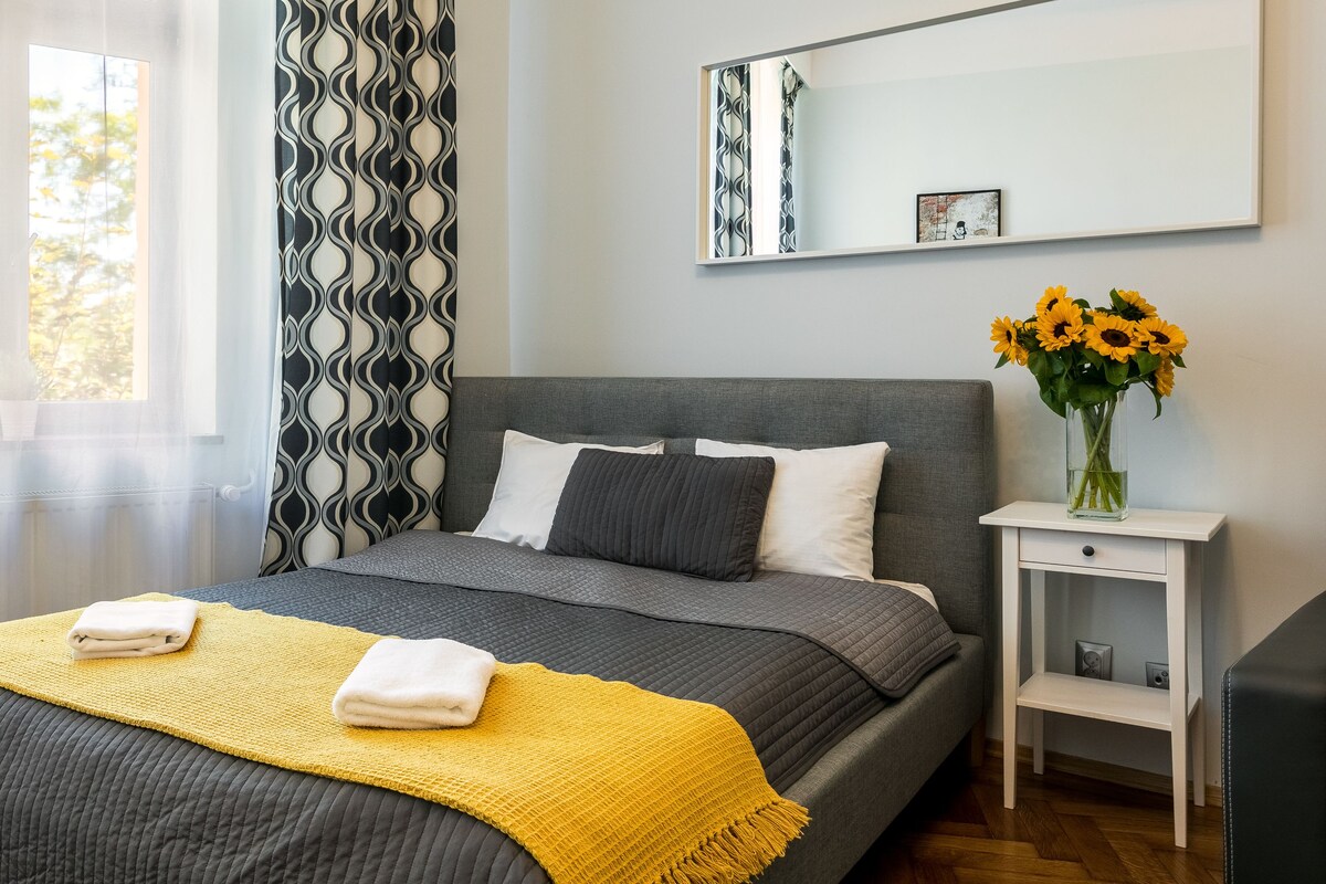 A modern bedroom with a grey bed, white pillows, a yellow throw, and a vase of sunflowers on a nightstand.