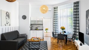 A modern living room with a black sofa, a coffee table, and a dining area with a table and chairs. There is a kitchen in the background with white cabinets and a hanging light fixture. - Old Town Residence BIS (Kraków)