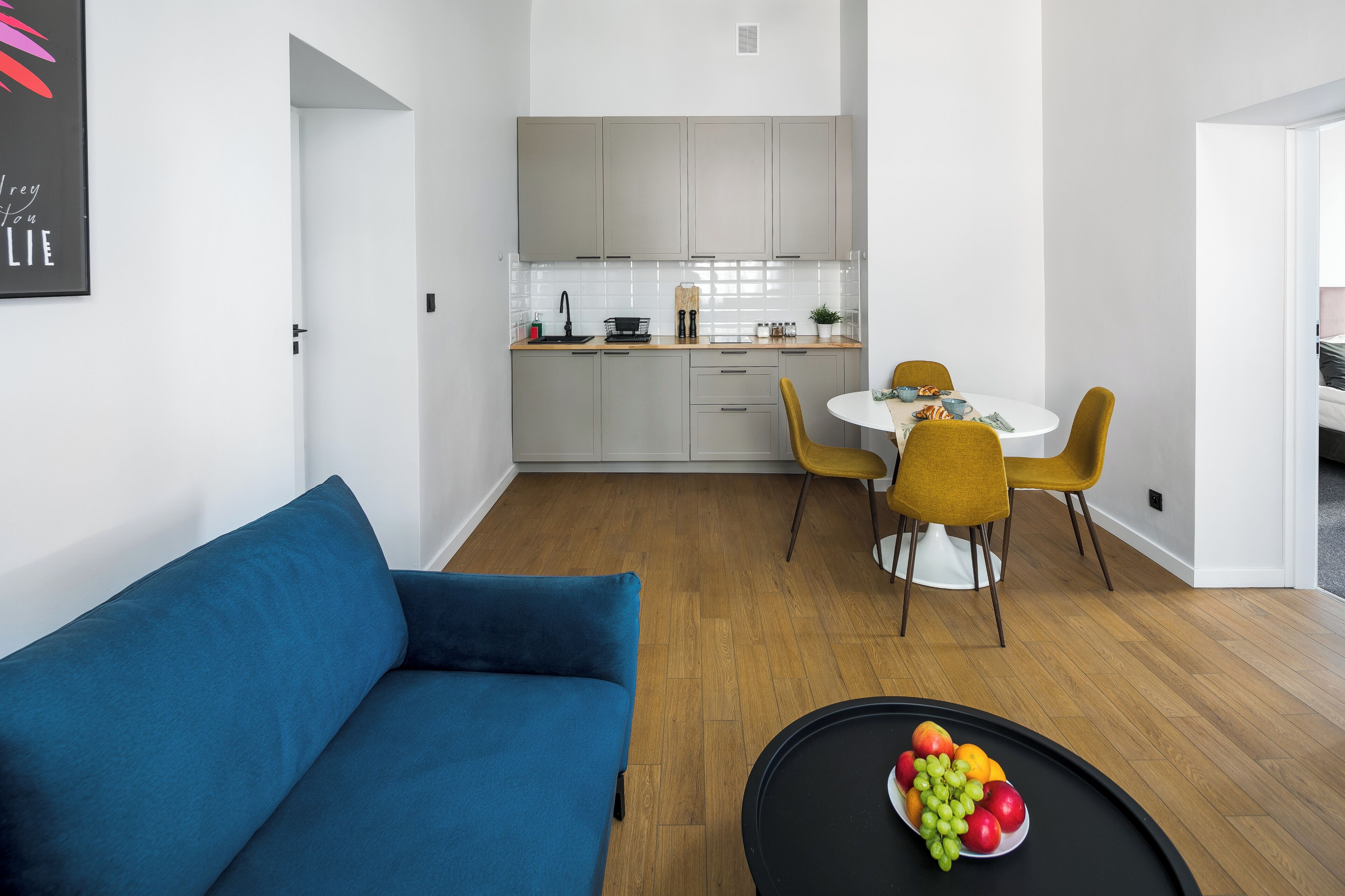 A modern living room with a blue sofa, a dining area with yellow chairs, and a kitchen with white cabinets.