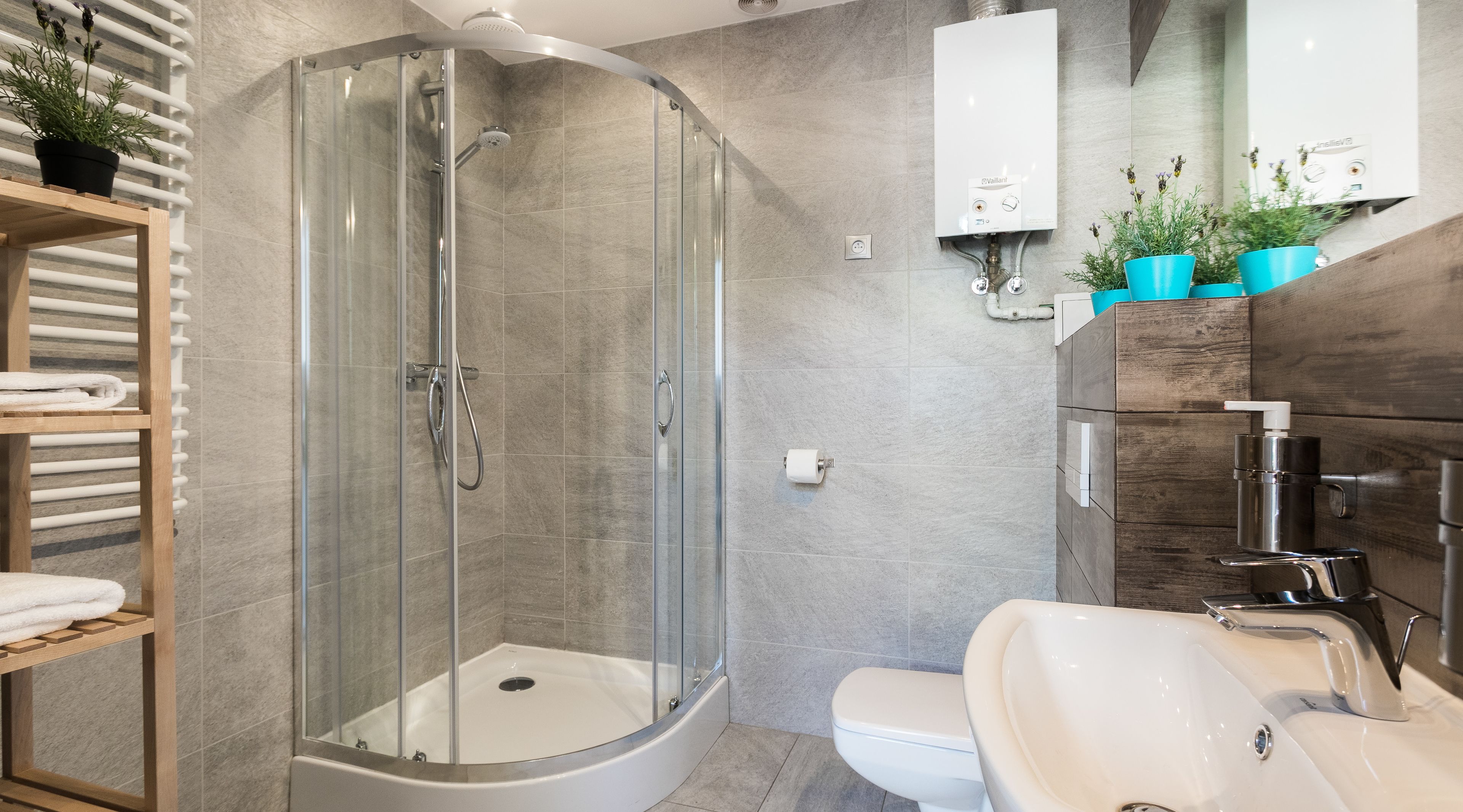 Une salle de bain moderne avec une douche à parois de verre, un lavabo blanc et des toilettes. Des plantes en pot sont disposées sur le mur, et une étagère en bois contient des serviettes.