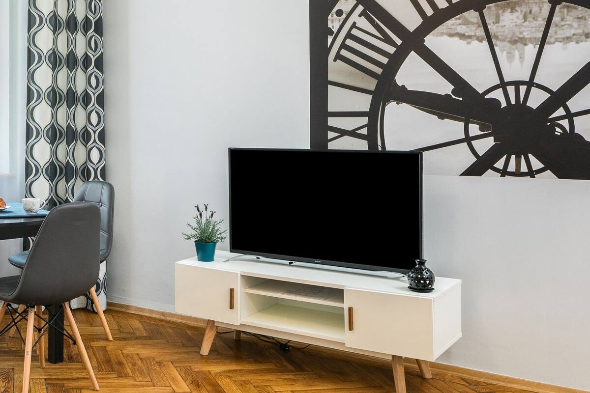 Un coin repas avec une table ronde, quatre chaises, un vase rempli de tournesols et un téléviseur posé sur un meuble blanc.