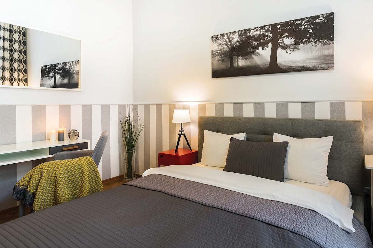 Un dormitorio ordenado con cama, mesita de noche, lámpara y un cuadro colgado en la pared.