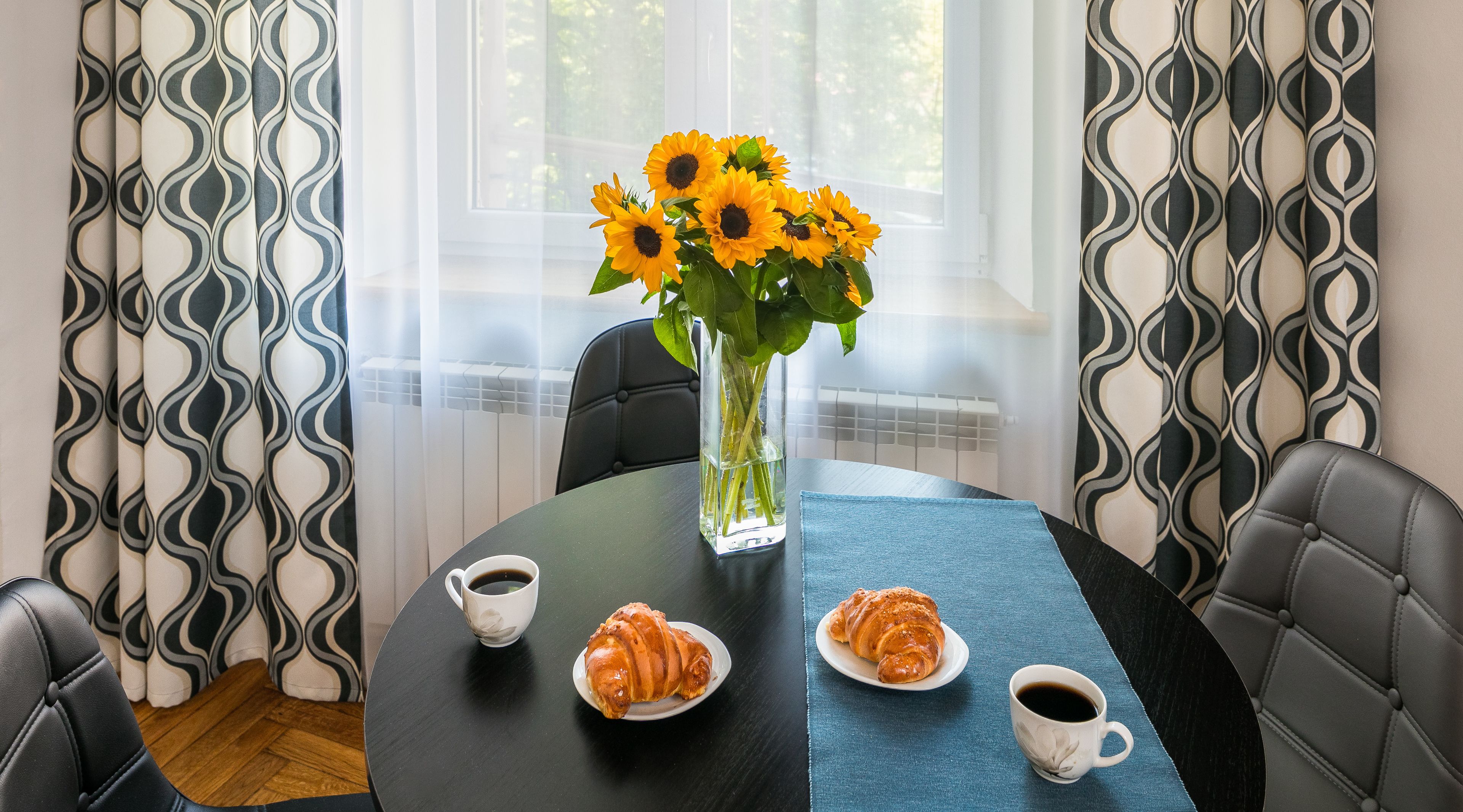 A dining table with a vase of sunflowers, two croissants, and two cups of coffee.