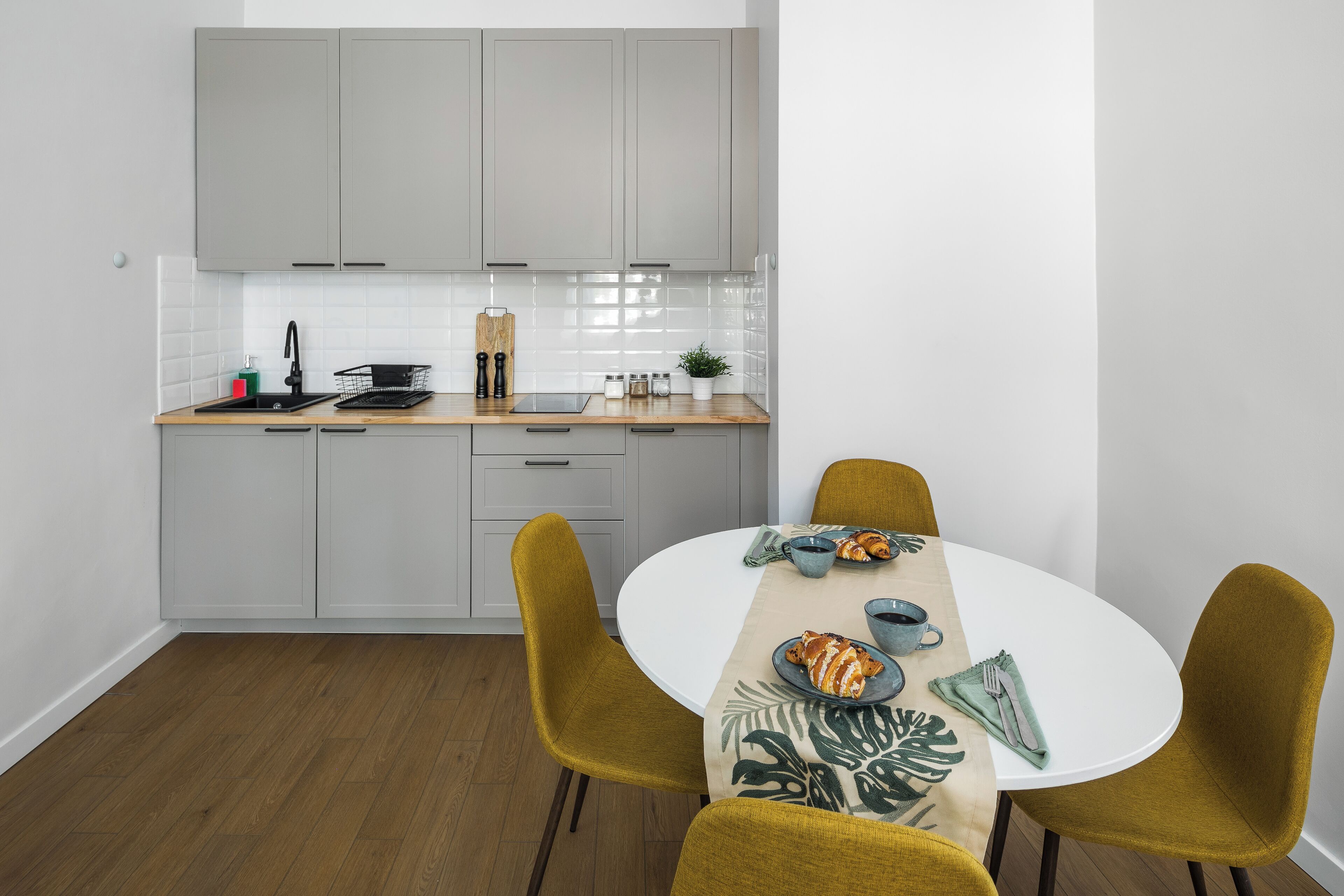 A modern kitchen with grey cabinets, a white countertop, and a dining area with a round table and mustard-colored chairs.