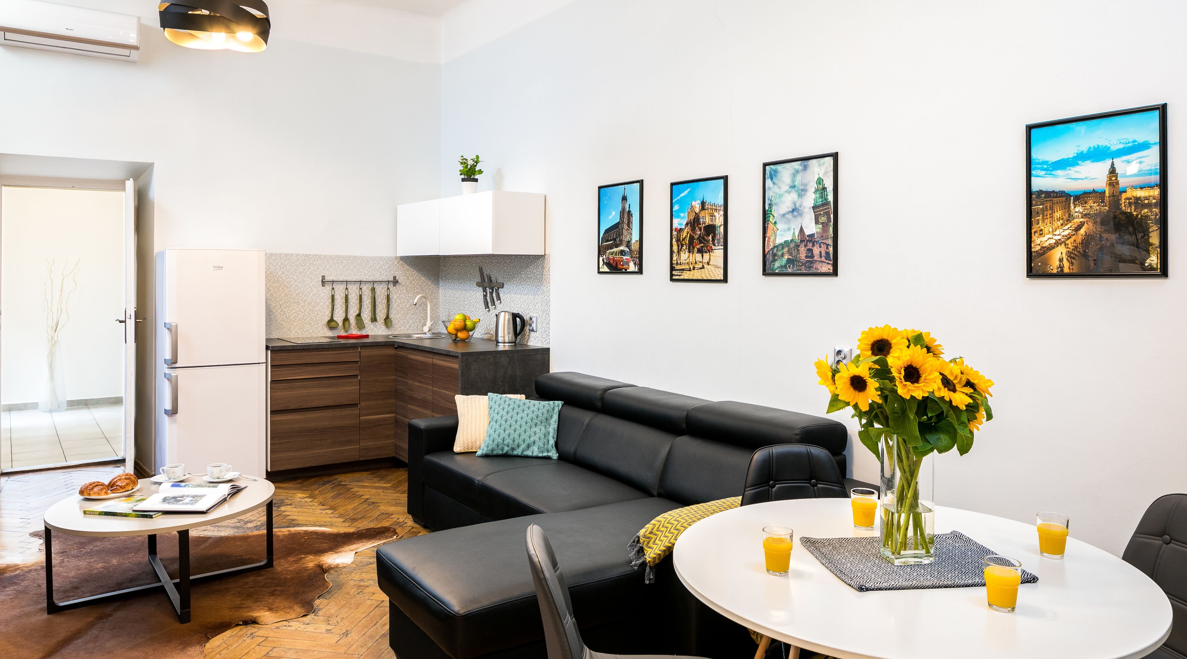 Un salon moderne avec un canapé en cuir noir, une table à manger avec deux verres de jus d’orange, et un coin cuisine avec un réfrigérateur blanc et un évier.
