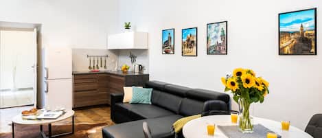 Un salon moderne avec un canapé en cuir noir, une table à manger avec deux verres de jus d’orange, et un coin cuisine avec un réfrigérateur blanc et un évier.