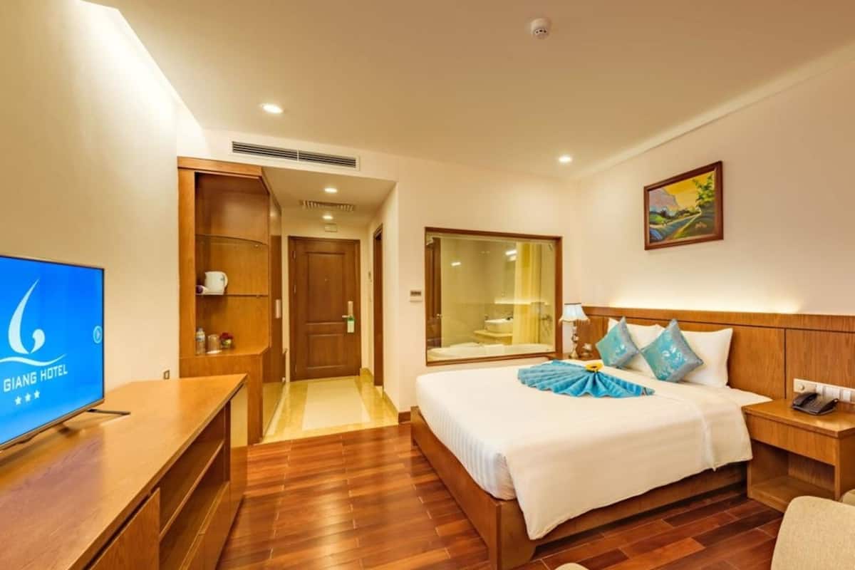 A hotel room with a wooden floor, a bed with white bedding, a bedside table, a nightstand, a TV, a bathroom with a glass shower enclosure, and a painting on the wall.
