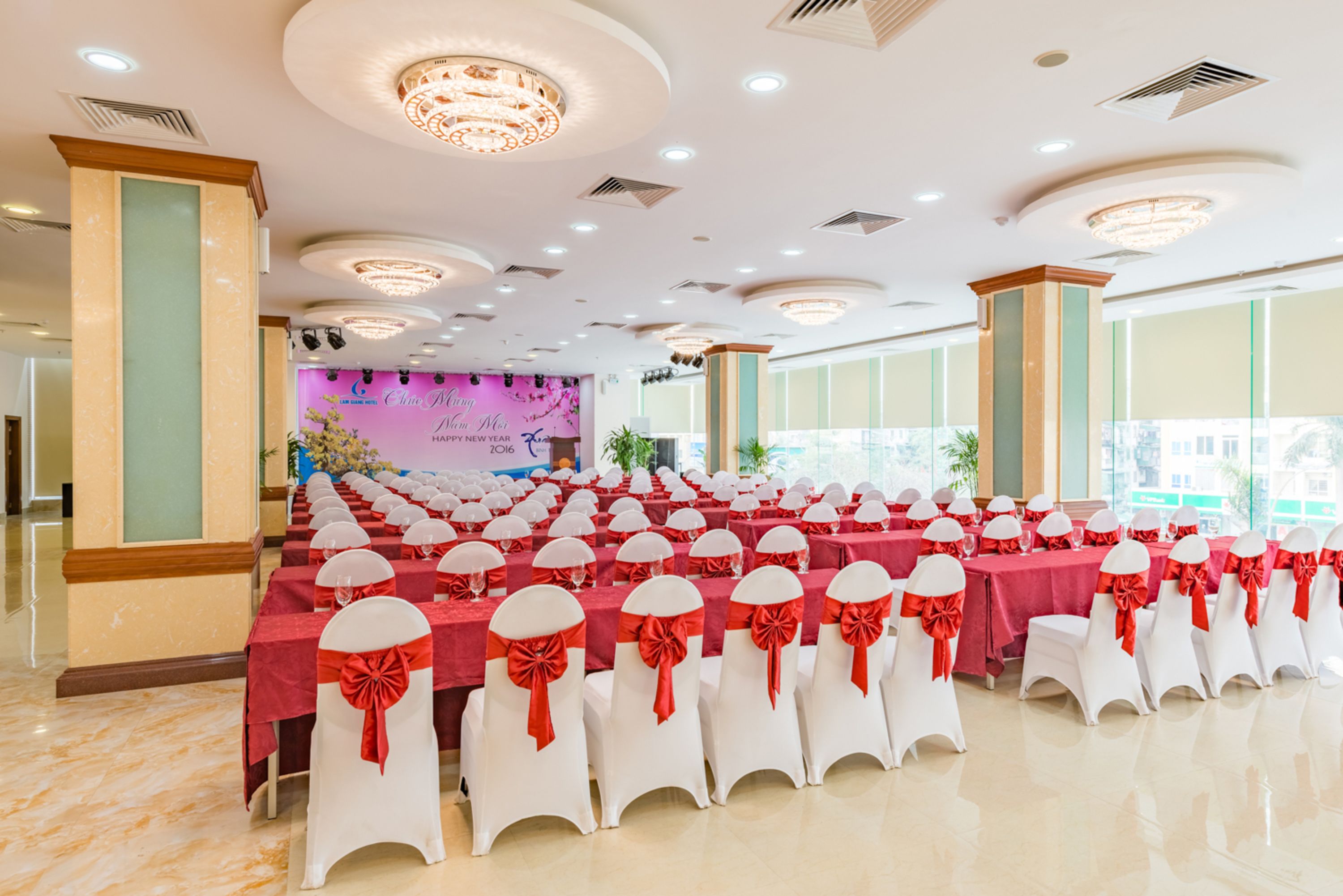 Uma sala de banquetes com fileiras de mesas e cadeiras, janelas amplas e um palco com um banner.