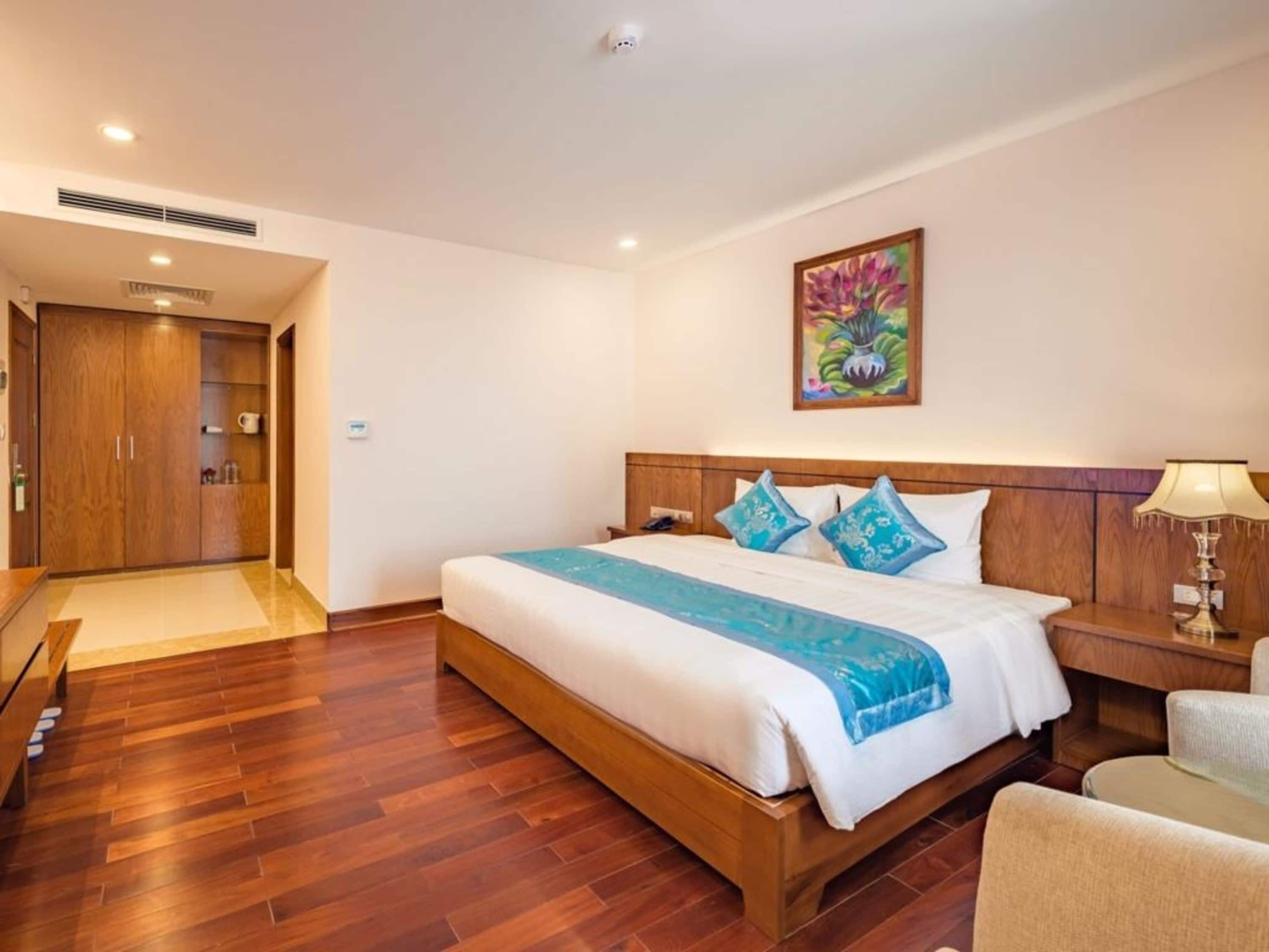 A hotel room with a wooden bed, a nightstand, a chair, a painting on the wall, and a wooden wardrobe.