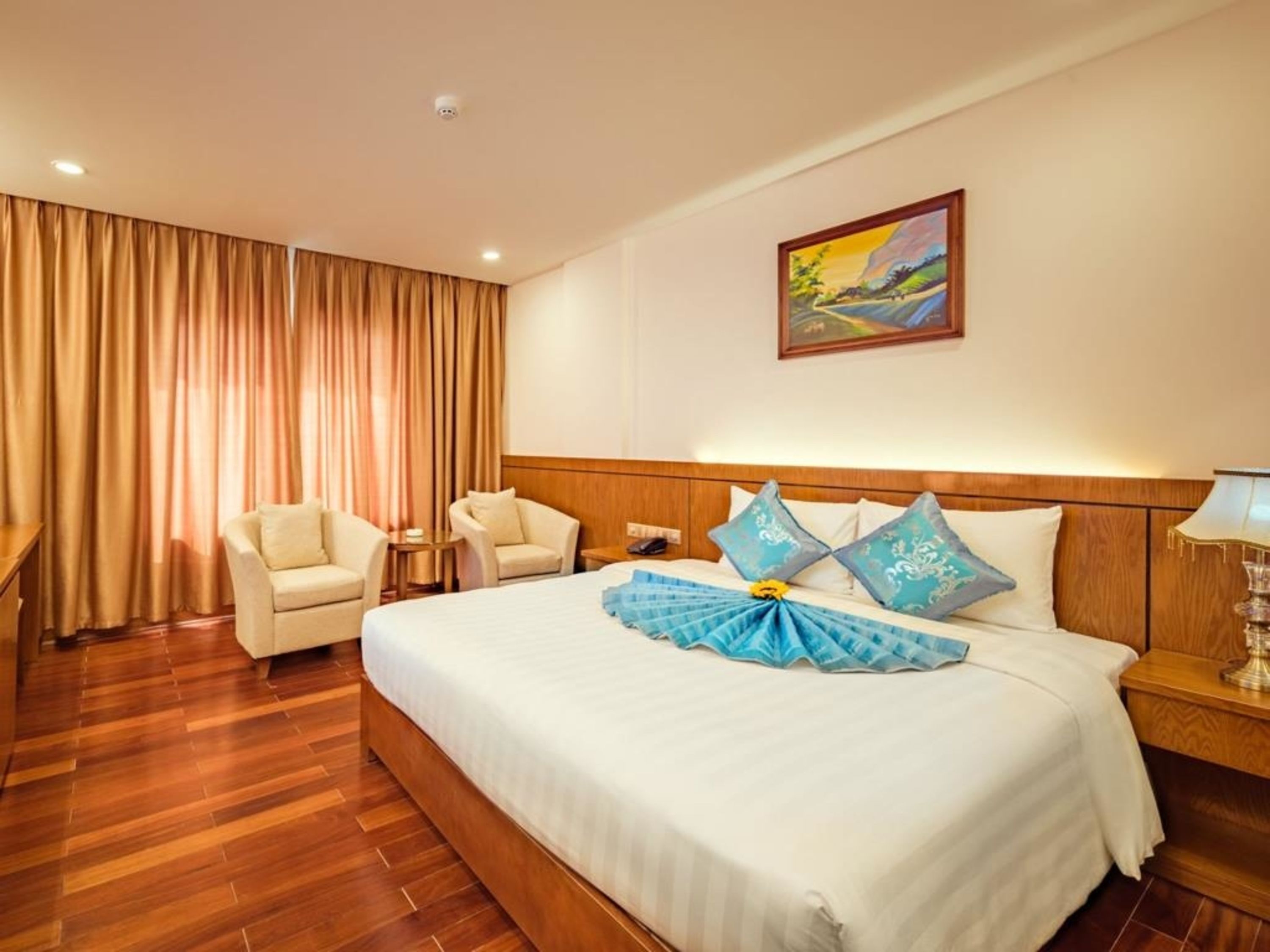 A hotel room with a large bed, two armchairs, a wooden headboard, a painting on the wall, and a curtain.