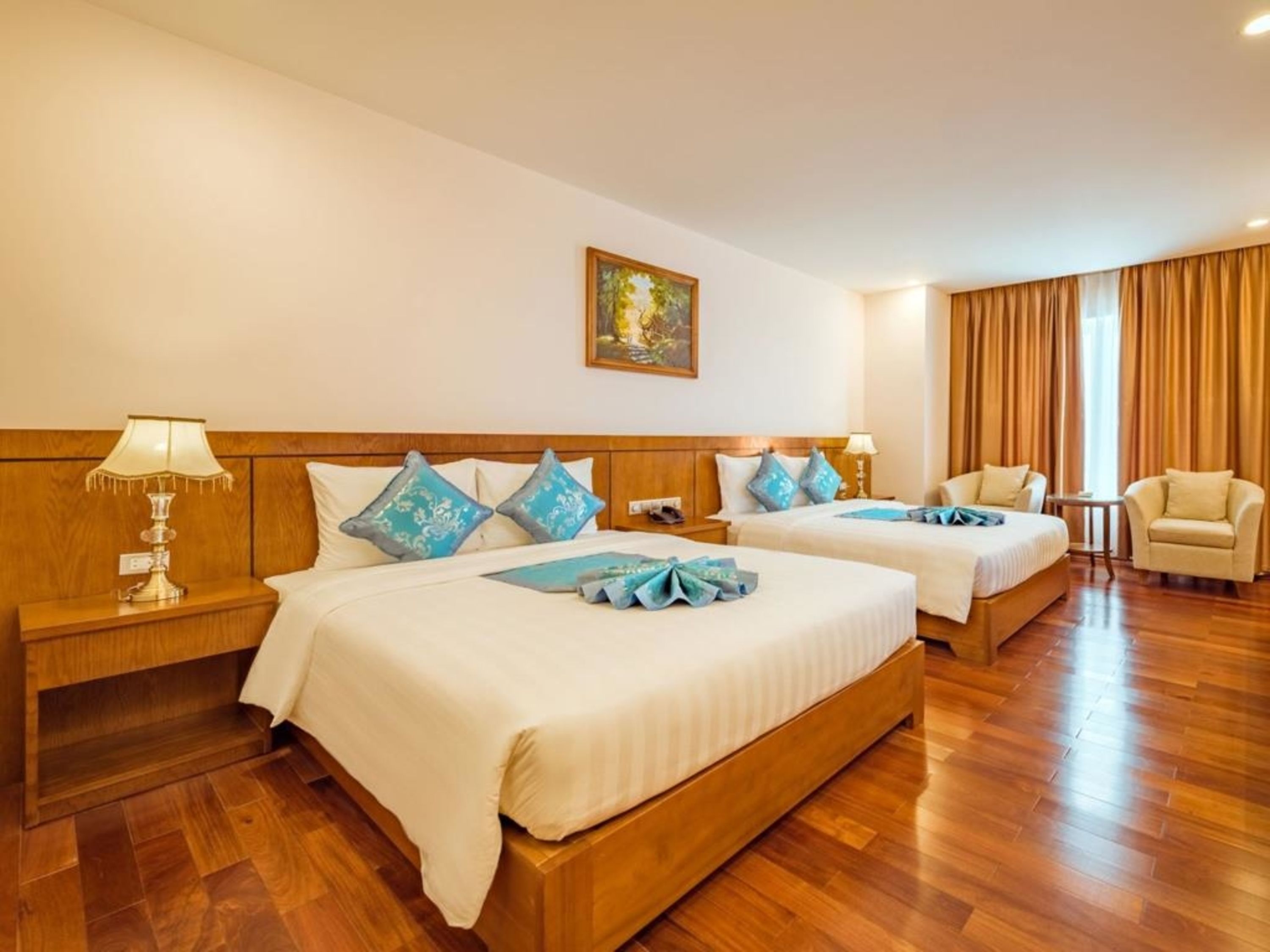 A hotel room with two beds, wooden headboards, a painting on the wall, a desk with a lamp, and a chair.