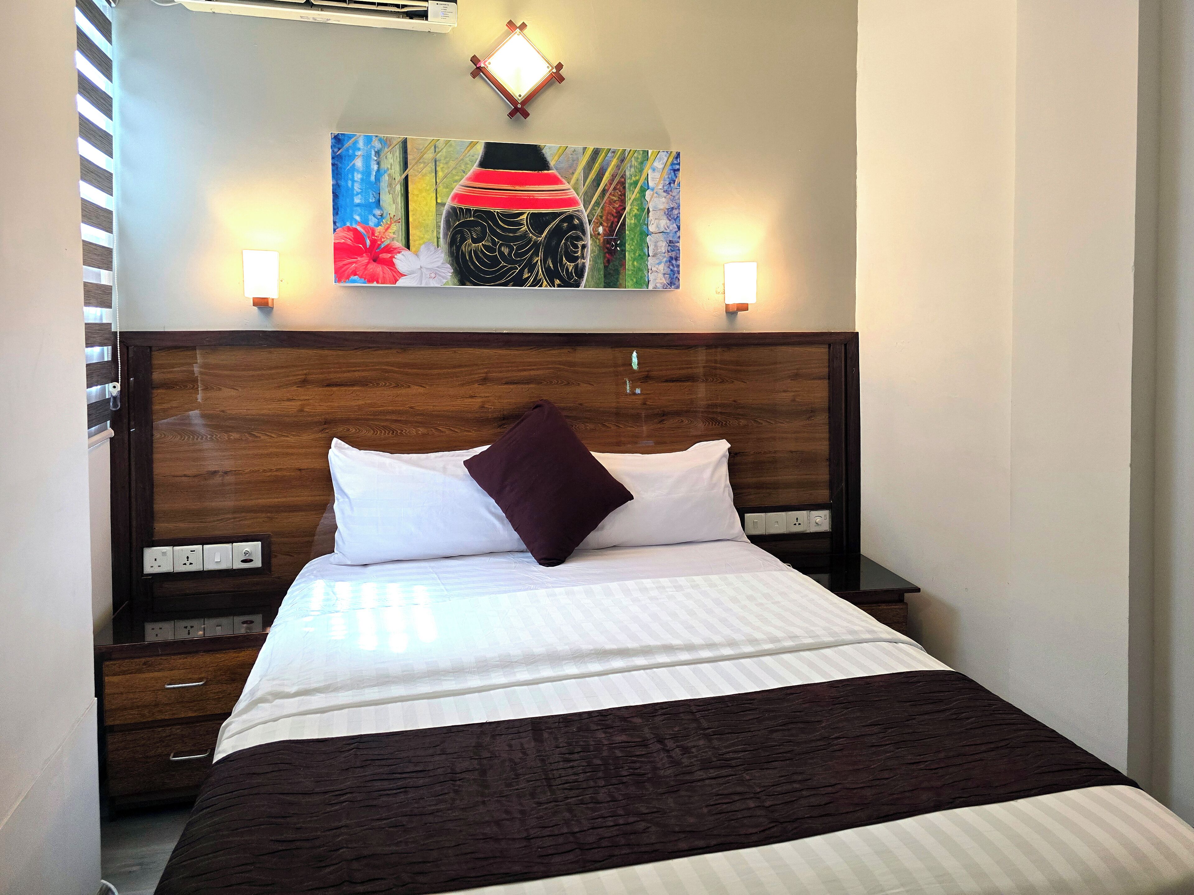 Habitación de hotel con cabecera de madera, cama con ropa de cama blanca y marrón oscuro, almohada morada y un cuadro sobre la cama.