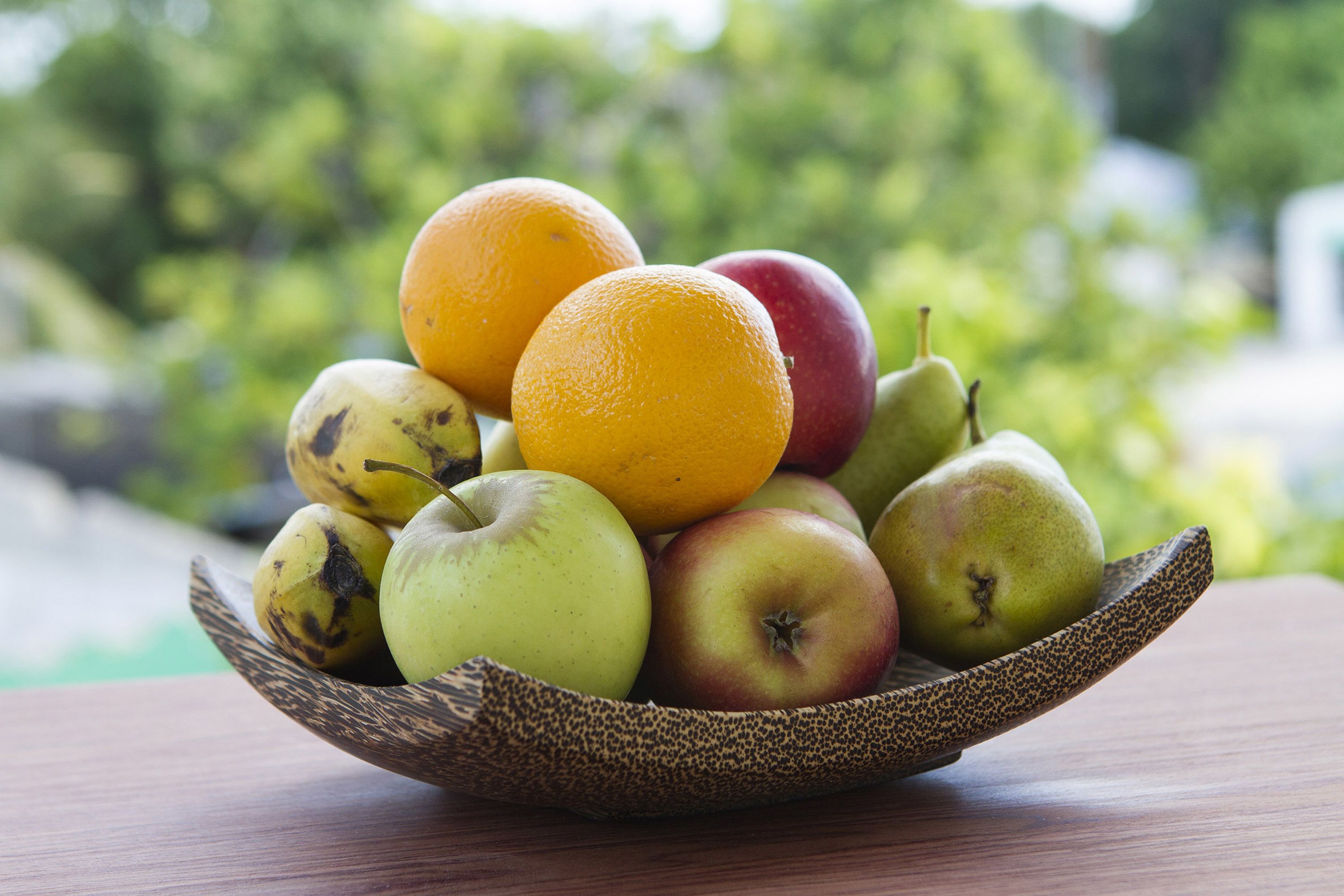 Un saladier rempli de fruits, notamment des pommes, des oranges et des poires, est posé sur une table en bois, avec un arrière-plan vert flou.