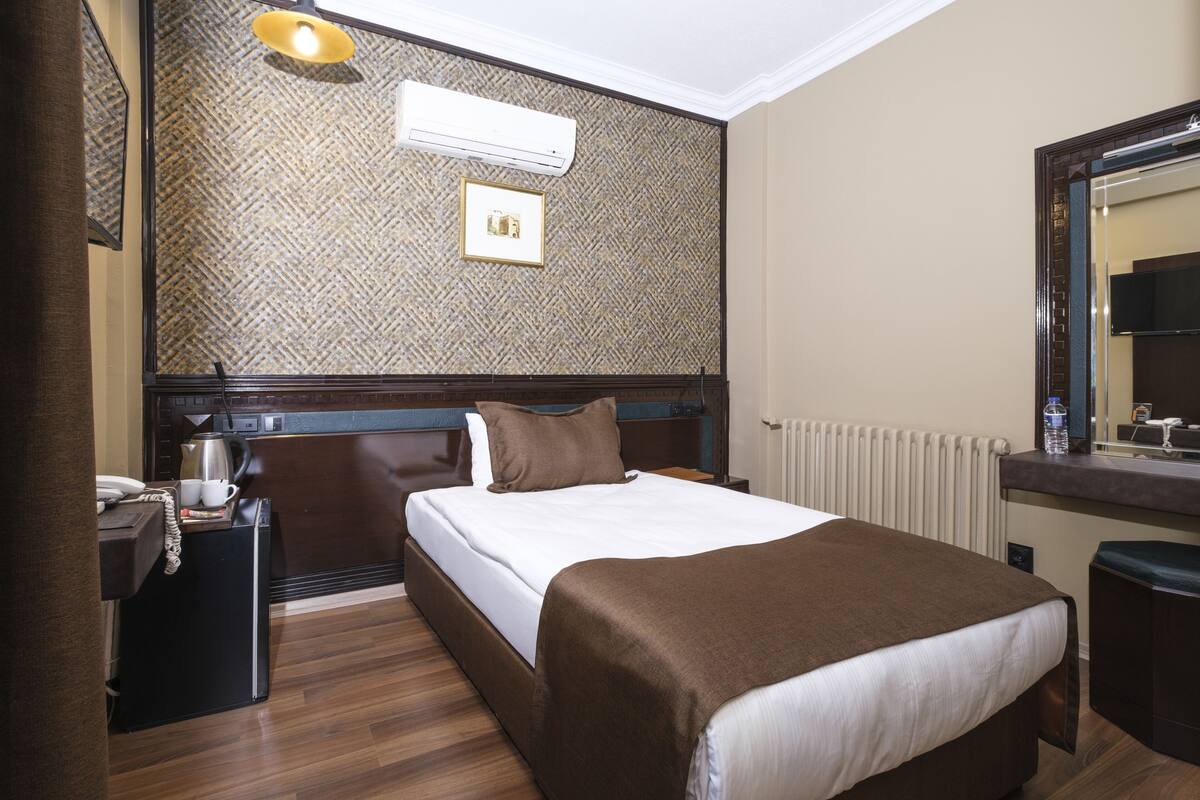 A hotel room with a wooden headboard, a bed with a white mattress and brown blanket, a bedside table with a lamp, a small refrigerator, and a mirror above the sink.