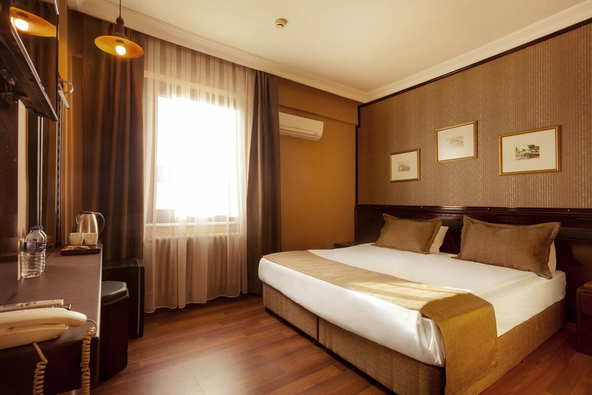A hotel room with a large bed, a desk with a telephone, a radiator, and framed pictures on the wall.