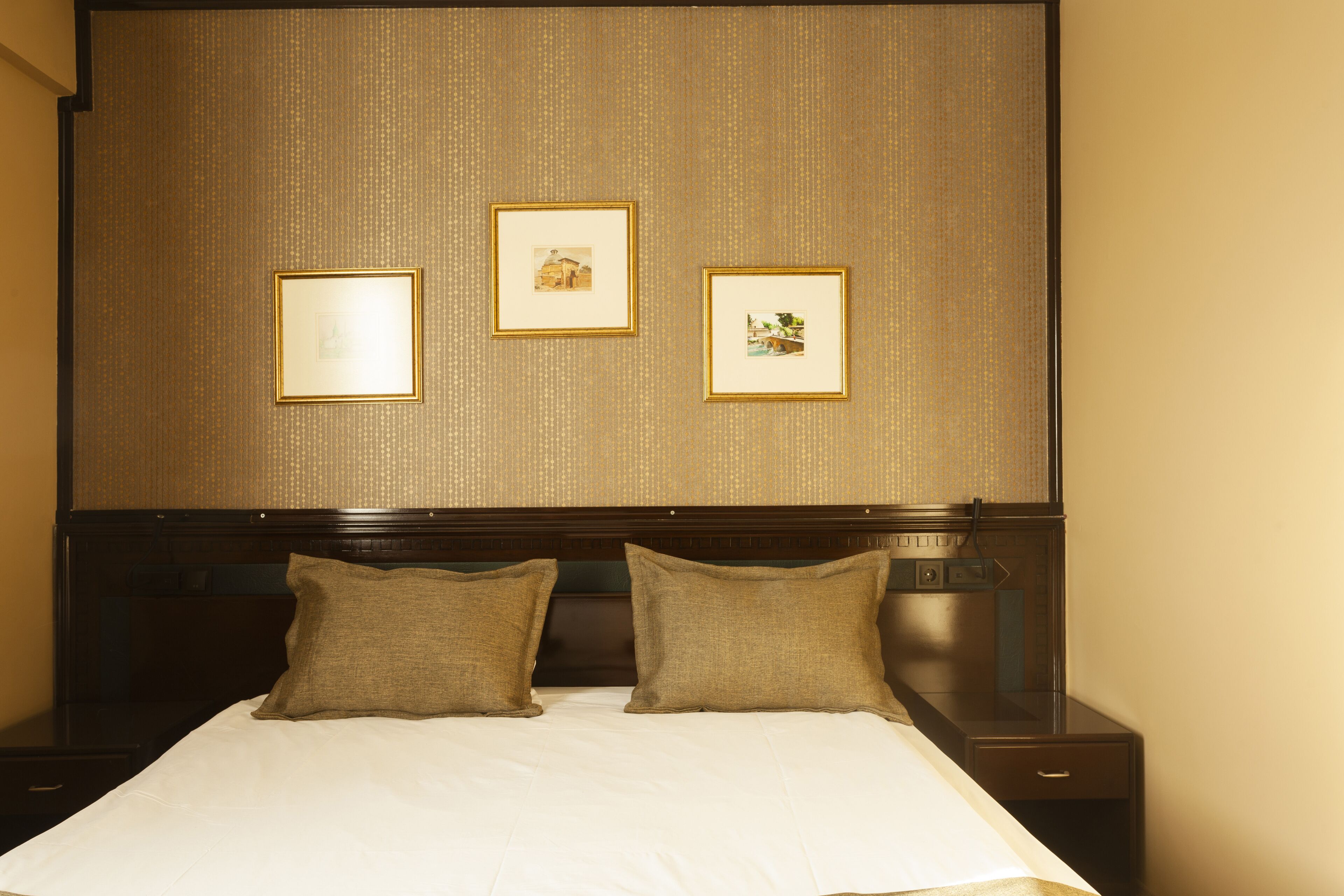 A hotel room with a large bed, two pillows, a headboard, and three framed pictures on the wall.