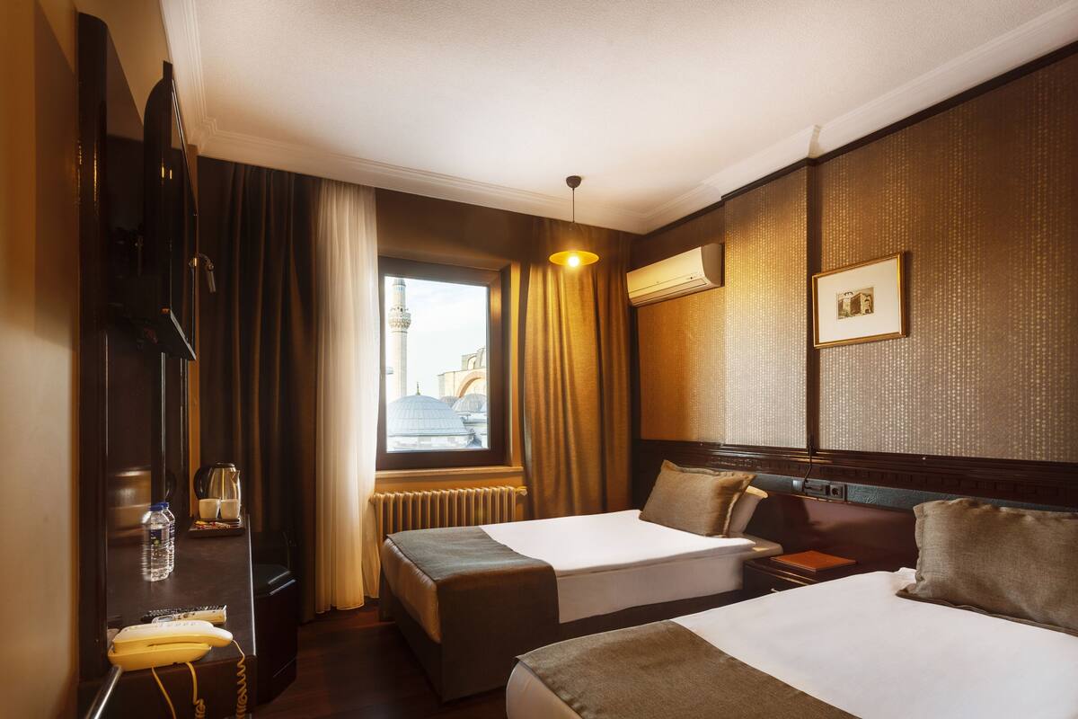 A hotel room with two beds, a desk with a telephone, a radiator, and a window with curtains.