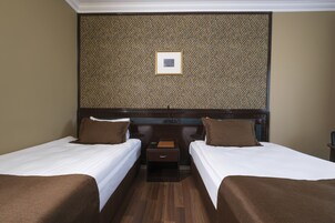 Habitación de hotel con dos camas individuales, cabecero de madera, mesita de noche pequeña con lámpara y un cuadro enmarcado en la pared.
