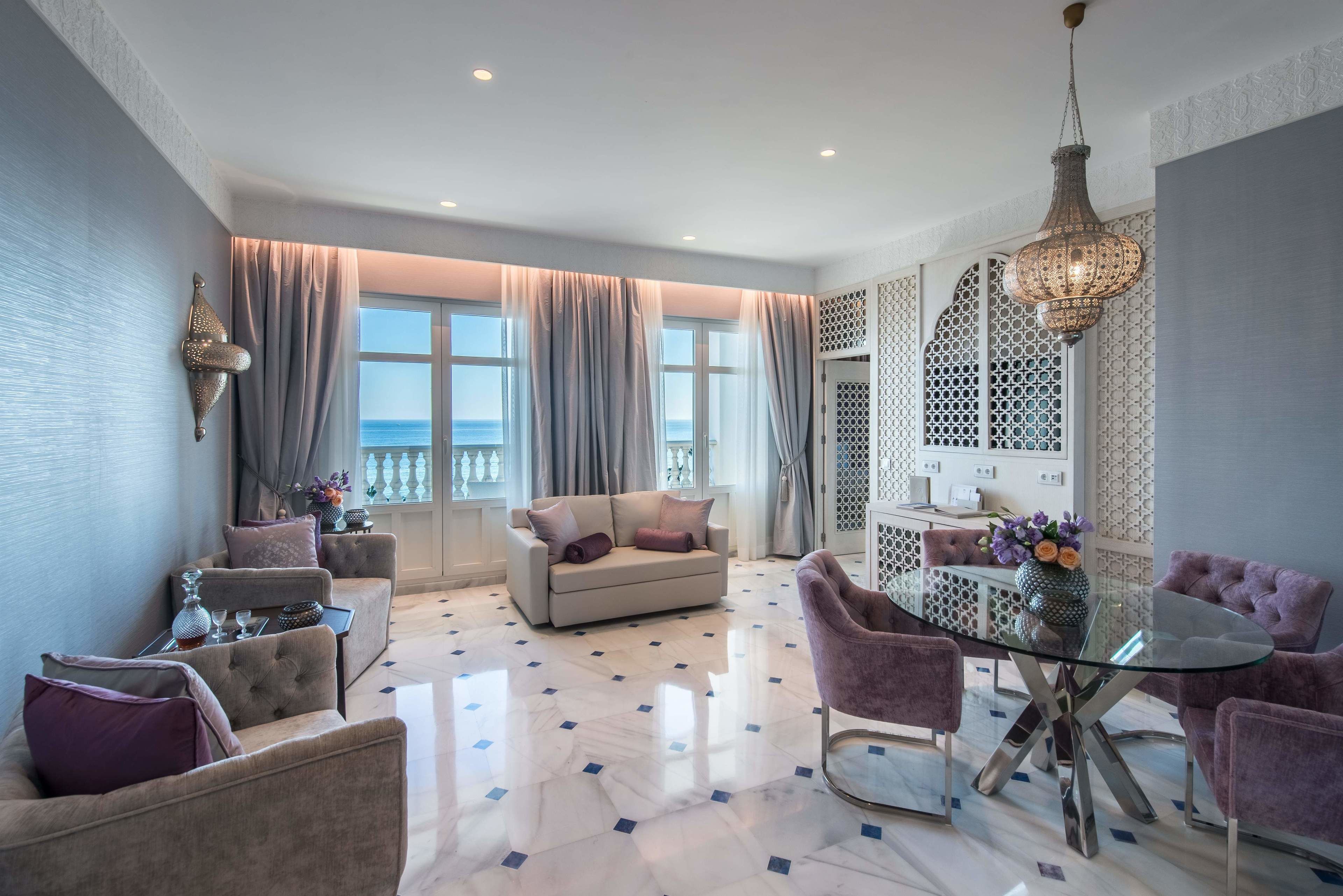 A modern living room with a sofa, armchairs, a glass dining table, and a view of the sea through large windows.
