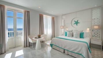 Una habitación de hotel moderna con una cama grande, una mesita redonda con un florero, una silla y vistas al mar a través de amplios ventanales.