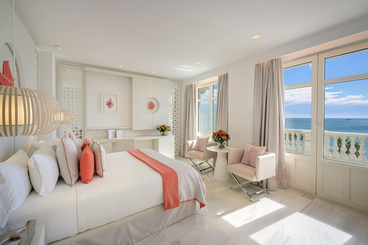 Habitación de hotel con una cama grande, un área de descanso con una mesa y sillas, y un balcón con vista al mar.