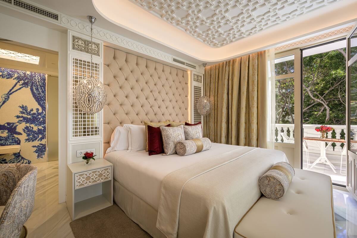 Un dormitorio con cabecera capitonada, un ventanal con vistas a árboles, una mesita de noche con un florero y una alfombra estampada.