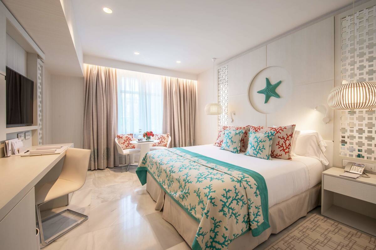 Una habitación de hotel moderna con cama, escritorio y silla, un ventanal con cortinas y un adorno de estrellas colgado en la pared.