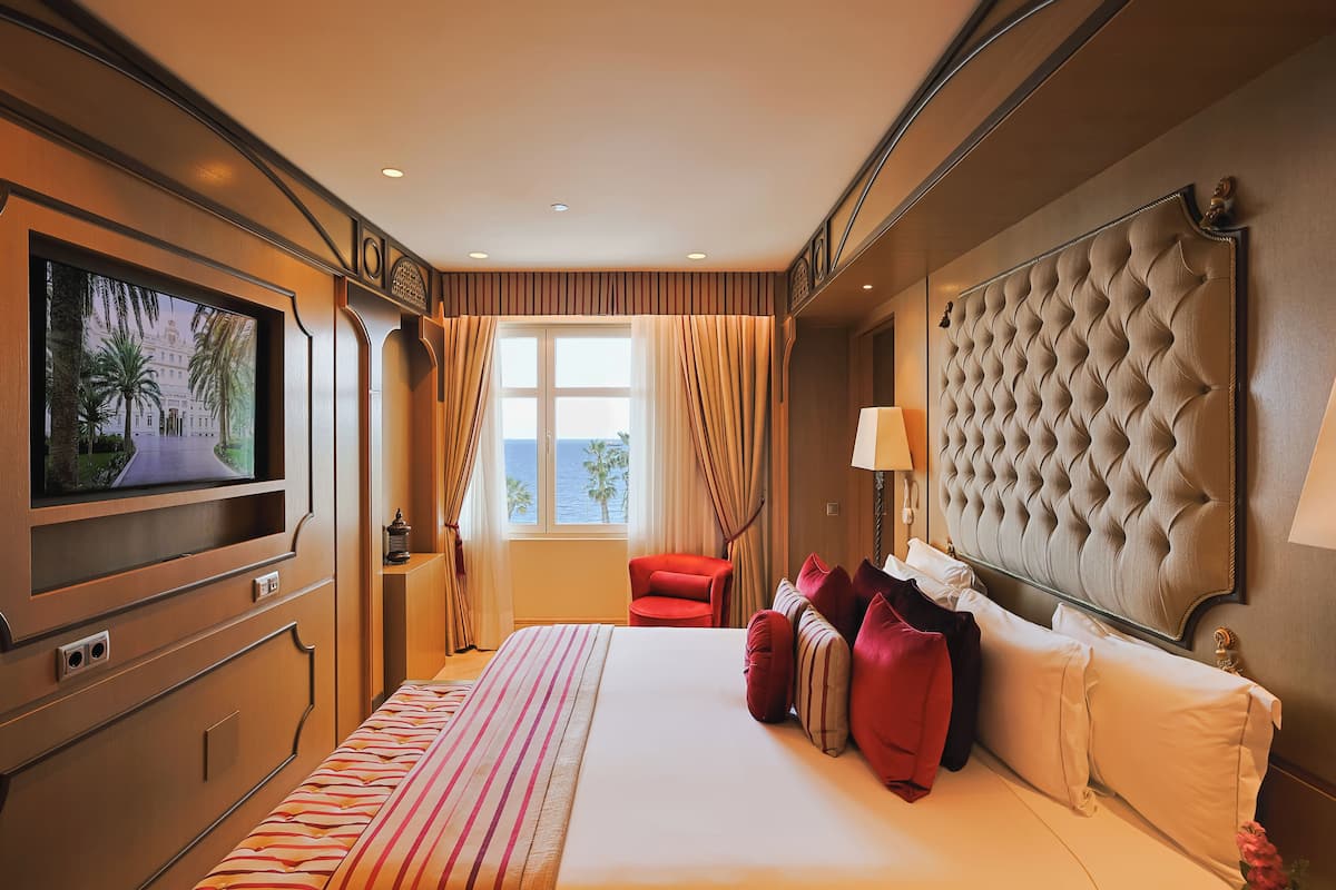 Una habitación de hotel con una cama grande, televisión, ventana con cortinas, una silla roja y un cabecero capitonado.