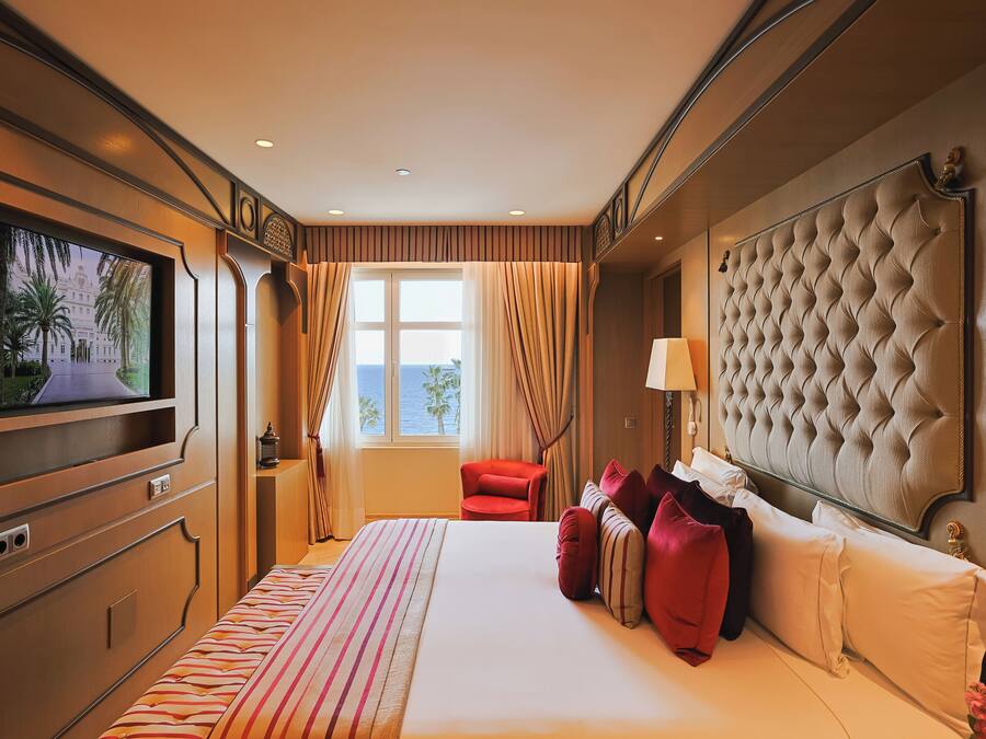 A hotel room with a large bed, a television, a window with curtains, a red chair, and a tufted headboard.