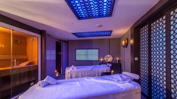 A modern spa room with two massage beds, a glass-enclosed shower area, and a ceiling with blue lighting.