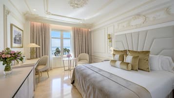 Un dormitorio lujoso con una cama grande, un escritorio y una silla. Se ve una ventana con cortinas, un jarrón de flores y un cuadro en la pared.