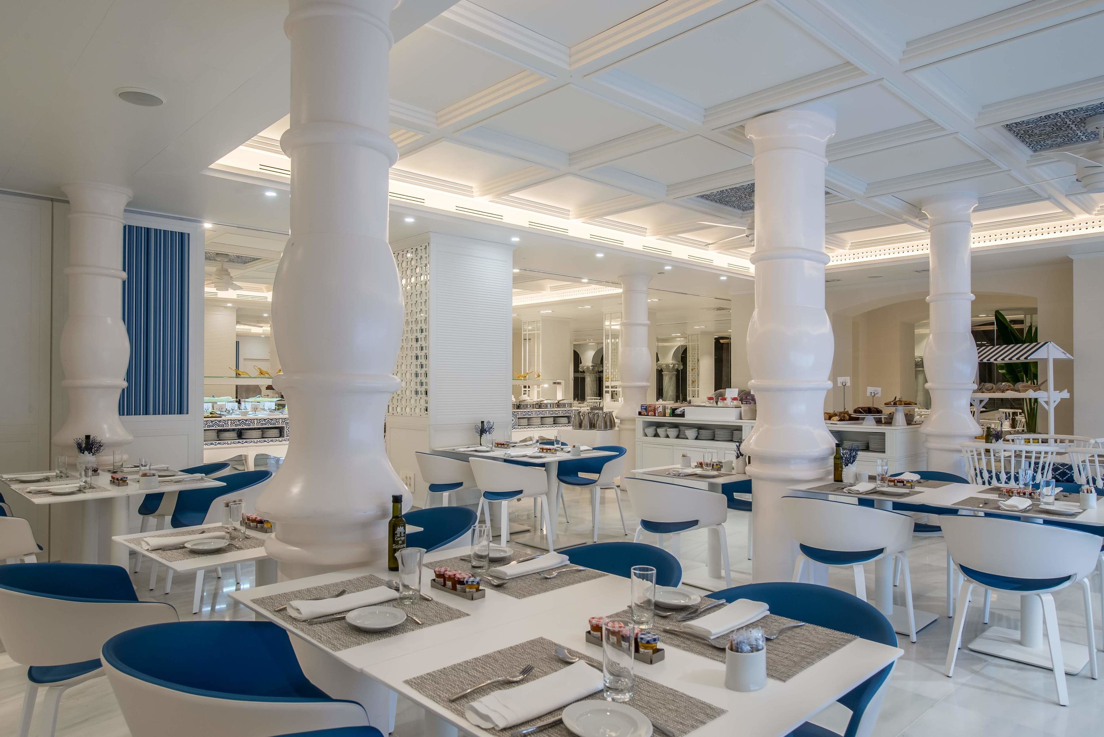 A spacious restaurant with white and blue chairs, large white columns, and a modern ceiling design.