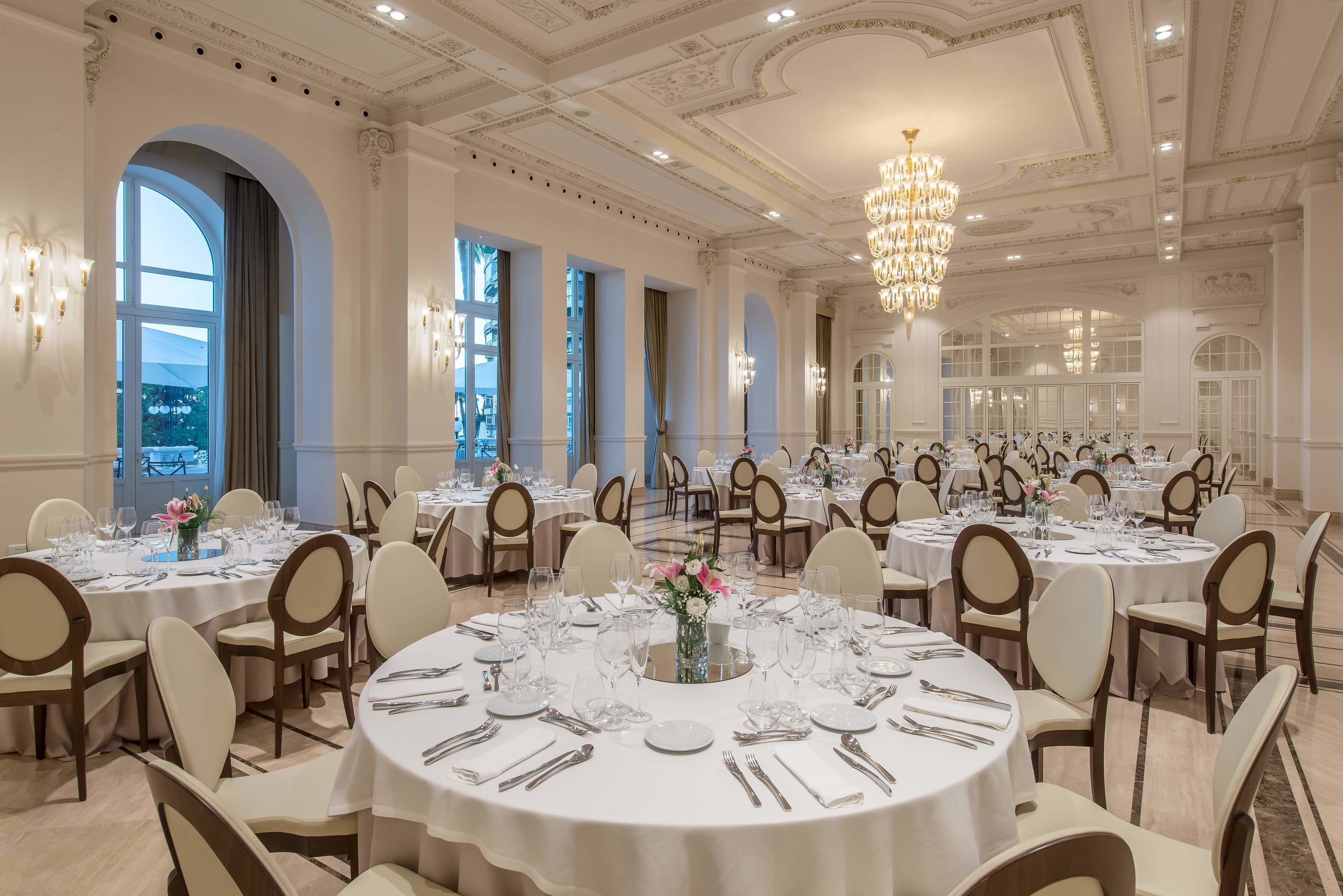 A spacious banquet hall with round tables, chandeliers, and elegant decor.