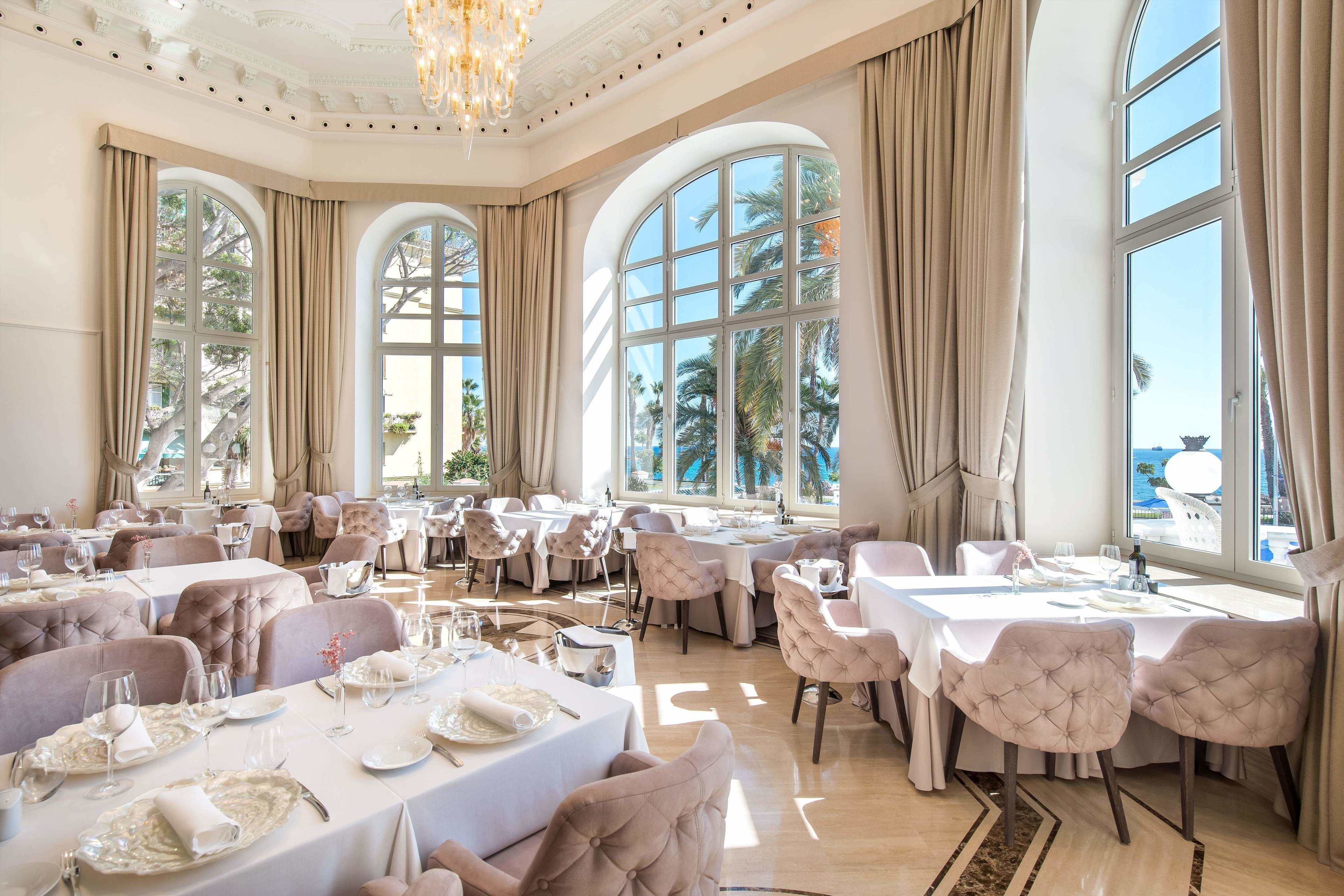 Un comedor amplio con varias mesas y sillas, ventanas grandes con cortinas y un candelabro.
