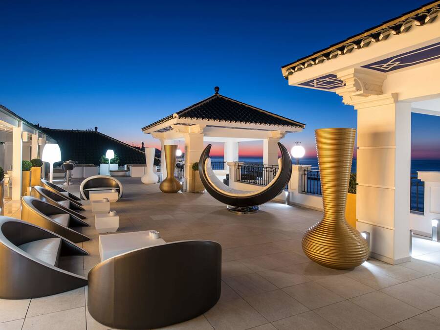 A rooftop terrace with modern seating, a decorative fountain, and a view of the sunset.