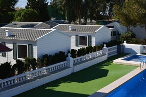 A row of white mobile homes with green shutters, a swimming pool, and a white fence with a decorative top. - Camping Valencia El Saler (Valencia)