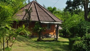 A small, single-story house with a sloped roof, surrounded by lush greenery and palm trees.