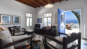Een woonkamer met een bank, fauteuils, een televisie en uitzicht op de zee door een open deur.
