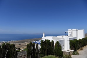 A white building with a checkered roof, surrounded by cypress trees, overlooking a coastal landscape. - Tinos Sky View Villa (Tinos)