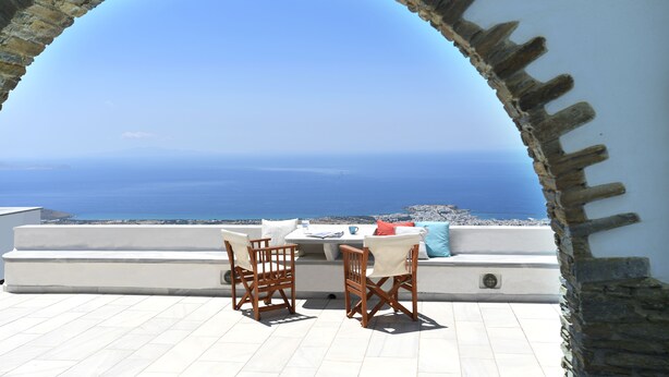 Een terras met een tafel en stoelen, met uitzicht op de kust door een rondboogvormige stenen constructie.