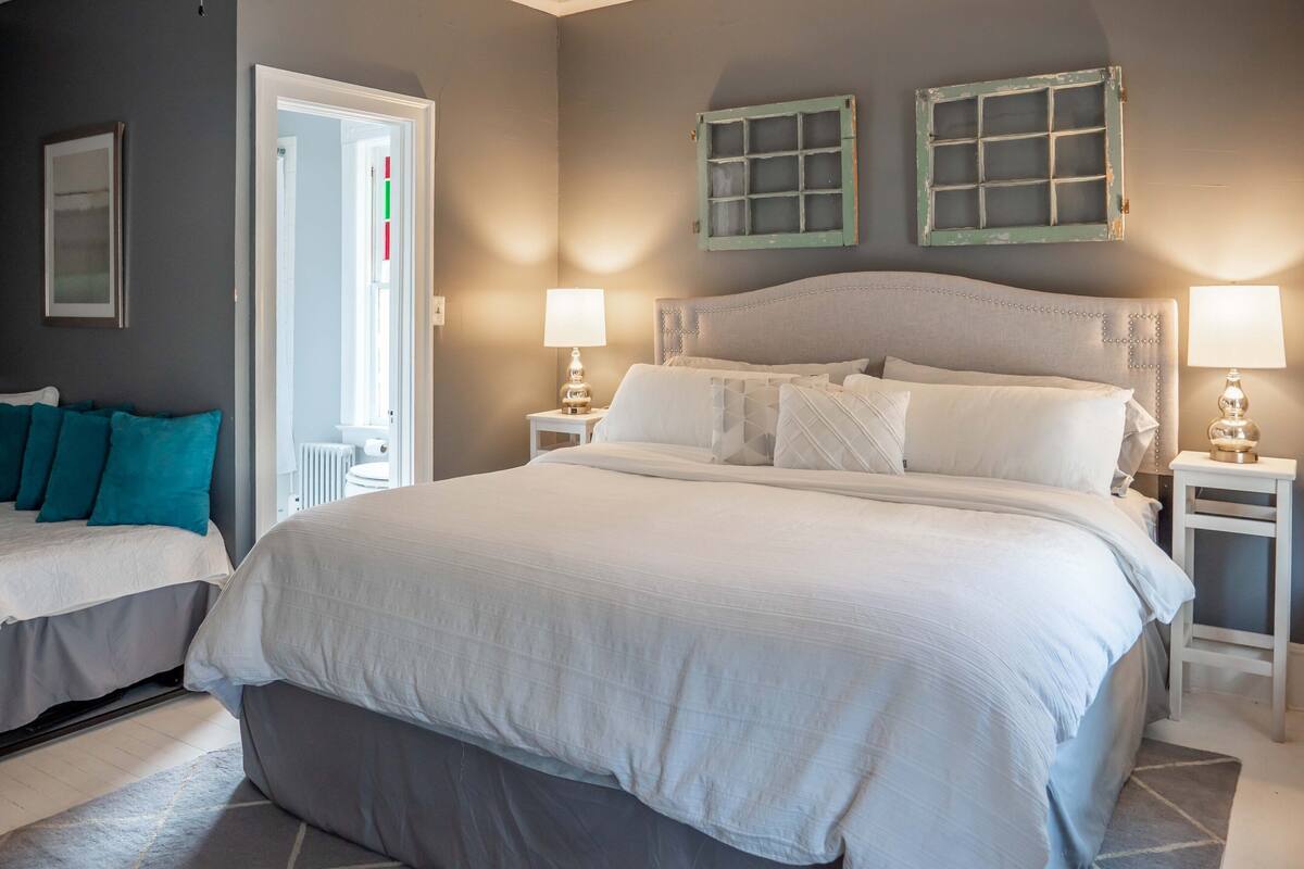 Un dormitorio con una cama grande, dos mesitas de noche, un buró, una puerta y una ventana con cortinas.